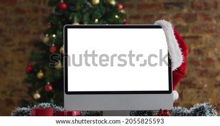 Santa hat hanging on computer monitor with copy space on screen, with christmas decorations and tree. christmas, festivity and communication technology digital composite image.