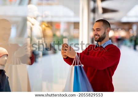 Happy millennial guy with shopper bags using smartphone at supermarket, taking selfie or photo, chatting with friend, copy space. Joyful young man checking mobile shopping list at mall