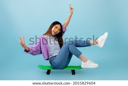 Joyful asian lady sitting on skateboard with legs up and smiling to camera, showing v-sign gesture. Young woman spending time and having fun over blue studio background
