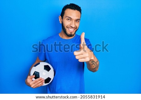 Hispanic man with beard holding soccer ball pointing fingers to camera with happy and funny face. good energy and vibes. 