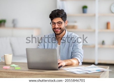 Portrait of happy arab freelancer man sitting at desk with laptop computer at home office, looking and smiling at screen, copy space. Young entrepreneur guy enjoying remote work Royalty-Free Stock Photo #2055846701