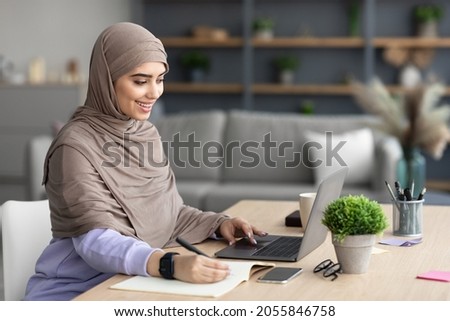 Note Taking. Portrait of positive arabian woman sitting at table, using laptop and writing in notebook, living room interior, copy space. Excited female student studying online from home, remote work