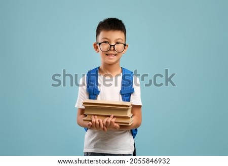 Funny Little Asian Schoolboy Wearing Eyeglasses And Backpack Holding Stack Of Books, Nerdy Korean Male Child Enjoying Study, Looking At Camera While Posing Over Blue Studio Background, Copy Space
