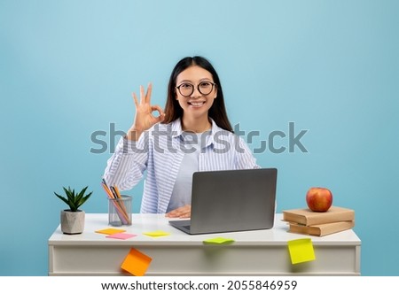 Happy asian female student in eyeglasses showing okay sign gesture sitting at the desk and using laptop over blue background. Lady doing homework, recommending online school or educational course Royalty-Free Stock Photo #2055846959