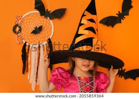 A girl on an orange studio background puts on a witch hat for Halloween, hiding her face under it. Cute shy kid. Halloween concept.