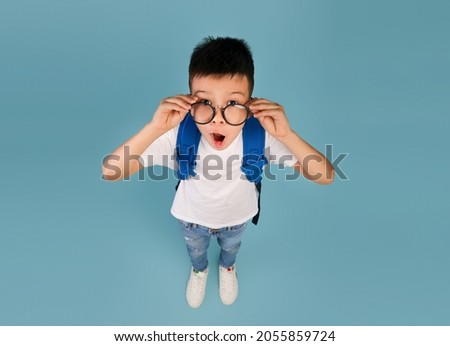 Back To School. Portrait Of Funny Excited Asian Schoolboy In Glasses, Surprised Korean Boy Carrying Backpack Posing Over Blue Studio Background And Looking At Camera, Top View With Copy Space