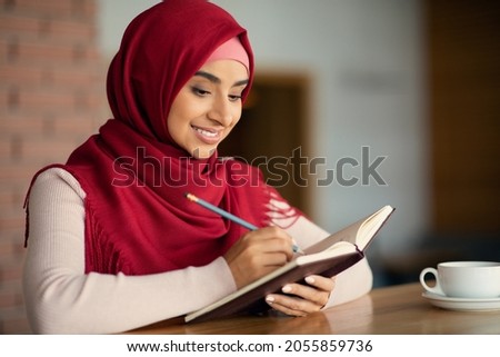 Smiling young muslim businesswoman in hijab writing notes while drinking coffe alone at cafe, planning her week while having lunch break, panorama with copy space, closeup portrait