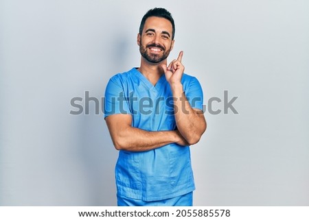 Handsome hispanic man with beard wearing blue male nurse uniform with a big smile on face, pointing with hand and finger to the side looking at the camera. 