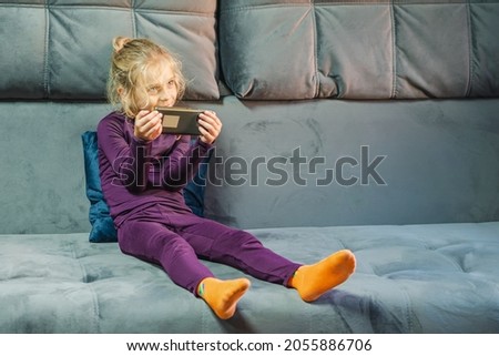 A blond girl in purple thermal underwear with a smartphone in her hands, sitting on a gray couch. The concept of a digital childhood