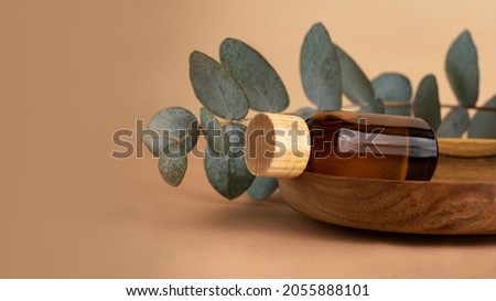 Cosmetics bottle from brown glass laying on the wooden plate with eucalyptus branch behind.Pastel background with fresh eucalyptus branch.Organic mockup concept,large banner.