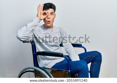 Young hispanic man sitting on wheelchair doing ok gesture shocked with surprised face, eye looking through fingers. unbelieving expression. 