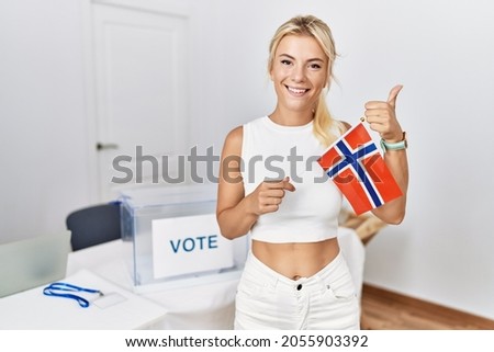 Young caucasian woman at political campaign election holding norway flag smiling happy and positive, thumb up doing excellent and approval sign 