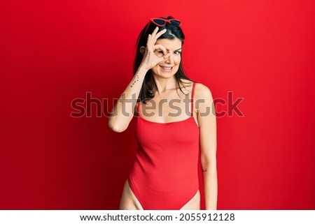 Young hispanic woman wearing swimsuit and sunglasses smiling happy doing ok sign with hand on eye looking through fingers 