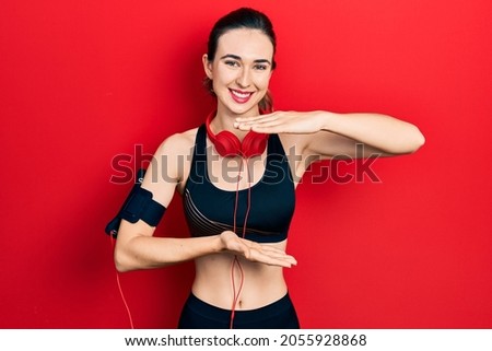 Young hispanic girl wearing gym clothes and using headphones gesturing with hands showing big and large size sign, measure symbol. smiling looking at the camera. measuring concept. 