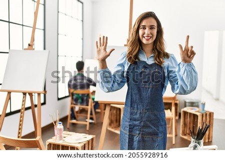 Young artist woman at art studio showing and pointing up with fingers number seven while smiling confident and happy. 