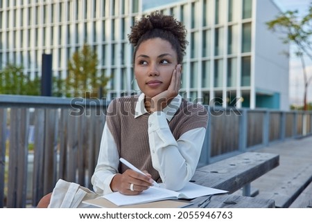 Thoughtful female student writes notes in notebook holds pen has pensive expression puts down her thoughts in personal diary during leisure time wears neat clothes makes schedule records checklist Royalty-Free Stock Photo #2055946763