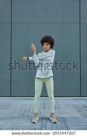 Vertical shot of sporty woman wears tracksuit stretches arms exercises outdoors has athletic body poses against grey wall focused forward has regular training. Athletic female warms up before running