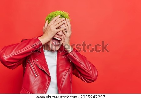 Horizontal shot of emotional hipster girl hides face with hands exclaims loudly keeps mouth widely opened wears leather jacket isolated over red background blank copy space for your advertisement