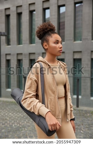 Motivated girl dressed in active wear goes for training carries rolled up yoga mat has fitness workout looks thoughtfully into distance poses in urban setting going to have morning stretching practice