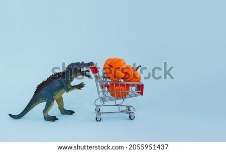 Small toy dino and shopping cart with decorative pumpkins on blue background. Halloween sale concept. Copy space.