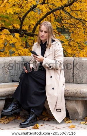 A blonde woman in an autumn park is sitting on a bench with a phone in her hands and a cup of coffee. Autumn leaf fall. People in the fall. Taking selfies, taking pictures on the phone,