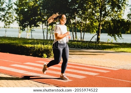 Full size portrait of a body positive plus-size plump woman athlete jogging running in fitness outfit, losing weight while listening to music in stadium outdoors Royalty-Free Stock Photo #2055974522