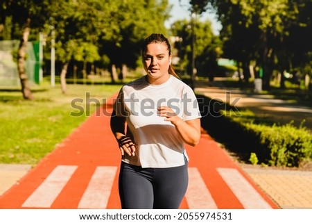 Body positive plus-size plump woman athlete jogging running in fitness outfit, losing weight while listening to music in stadium outdoors Royalty-Free Stock Photo #2055974531
