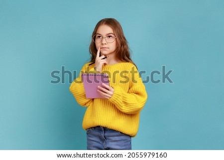 Portrait of little girl writing in notebook, thinking, taking notes, school child doing homework, wearing yellow casual style sweater. Indoor studio shot isolated on blue background. Royalty-Free Stock Photo #2055979160