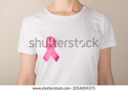 Photo of young woman in white t-shirt with pink ribbon symbol of breast cancer awareness on isolated grey background