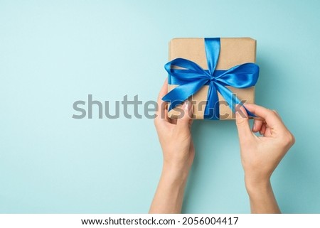 First person top view photo of hands unpacking craft paper giftbox with blue ribbon bow on isolated pastel blue background with copyspace