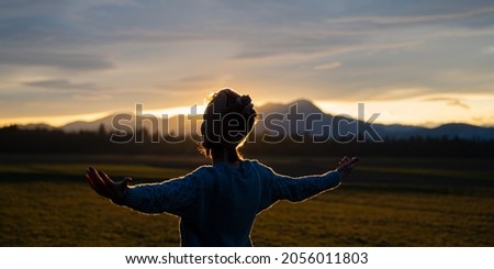 Young woman standing in beautiful nature at sunset meditating with arms spread gently lit by the setting sun. Royalty-Free Stock Photo #2056011803