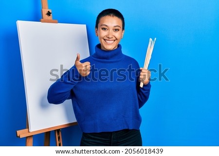 Beautiful hispanic woman with short hair holding brushes close to easel stand smiling happy and positive, thumb up doing excellent and approval sign 