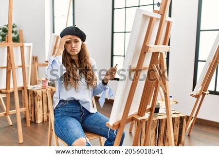 Young hispanic artist woman painting on canvas at art studio worried and stressed about a problem with hand on forehead, nervous and anxious for crisis 