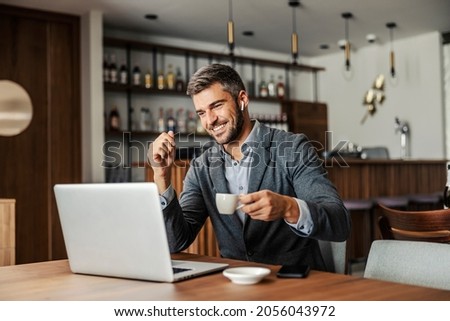 A happy businessman is sitting in a cafe, drinking coffee and watching a webinar on the laptop. The man has earphones in his ears so he can listen to the lecture. A businessman using laptop in cafe Royalty-Free Stock Photo #2056043972