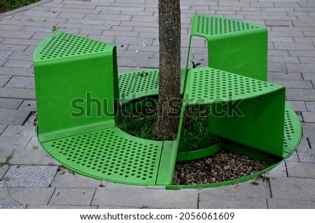 bench sitting with a hole around a tree. green painted metal perforated plate. three places around the tree around like a pizza or a shamrock. concrete pavement path in the park