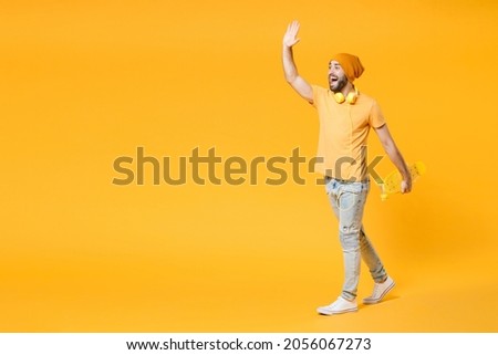 Full length side view of funny excited young man in basic casual t-shirt headphones hat waving and greeting with hand as notices someone hold skateboard isolated on yellow background, studio portrait