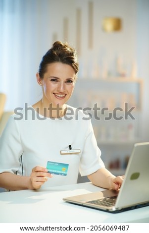 smiling female employee with credit card and laptop buying on internet in modern beauty salon.