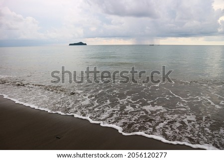 a beach with black sand and a small island in the middle of the sea, the picture was taken in the afternoon when the sky was a little cloudy