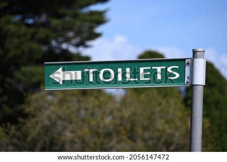 A green outdoor toilets sign with white lettering and arrow on a sunny day, with trees and blue sky in the background