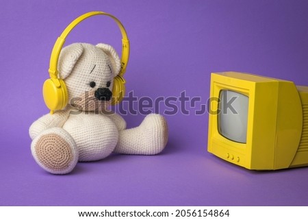 A white bear cub with headphones in front of the TV on a purple background. The concept of virtual dependence of children.