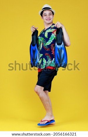 Young attractive Asian man wearing green and blue Hawaiian shirt holding blue flippers and going for beach vacation against yellow background.