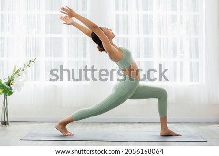 Side view of Asian woman wearing green sportwear doing Yoga exercise in front of windows,Yoga high lunge pose or Anjaneyasana,Calm of healthy young woman breathing and meditation yoga at home Royalty-Free Stock Photo #2056168064