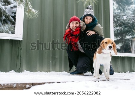 Adorable couple posing for a photo with their beagle outdoors at winter. Man is crouched, woman is leaning on him from behind. Dog is in front of them, facing the camera.