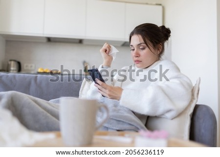 Sick young female with flu browsing, surching possible symptoms on internet by phone, consulting with doctor online on sofa at home. Ill girl with cold chatting with physician in medical app. Royalty-Free Stock Photo #2056236119