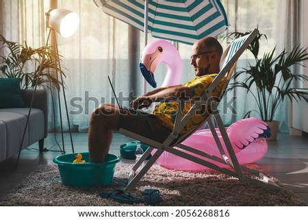 Man spending summer vacations at home alone, he is sitting on the deckchair in the living room and working with a laptop
