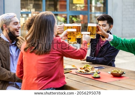 Multiracial friends with face mask under the chin cheering with large beer at brewery bar - Millennial hipster people having fun together drinking alcohol - Focus on beer glasses