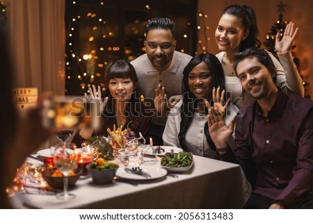 holidays and celebration concept - multiethnic group of happy friends photographing at christmas dinner and waving hand