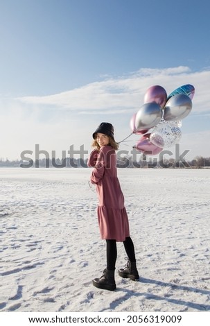 Portrait of tender teenage girl in pink dress, black panama hat, holding many multi-colored helium balloons in hands. sunny winter day. feels great, festive, enjoys moment. Beautiful birthday girl