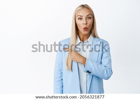 Surprised businesswoman with blond long hair, pointing left, gasping and say wow, impressed with announcement, white background
