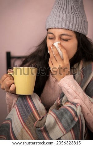 indoor image of a young woman getting sick with flu and cold and using a tissue paper to sneeze and blowing her nose while holding a coffee mug at home in winters. She is wearing warm clothes. Royalty-Free Stock Photo #2056323815